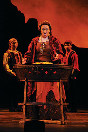 A woman in a red and brown costume stands behind a wooden table, passionately performing on stage with two similarly dressed people in the background and a dramatic orange backdrop.