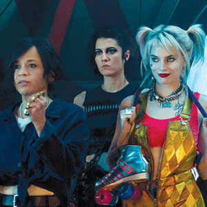 Three women stand together; the one in front left has short dark hair and a blue jacket, the middle woman has dark hair and a black top, and the right woman has blonde pigtails, a gold outfit, and holds roller skates.