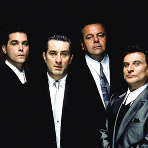 Four men in suits stand close together against a black background, looking seriously at the camera.
