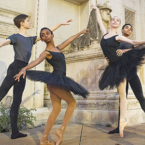 Four ballet dancers in black attire pose gracefully outdoors in front of a stone building with classical sculptures, each extending their arms and legs in elegant ballet positions.