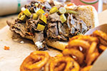 A sandwich filled with sliced beef and topped with chopped pickled vegetables is served on a piece of bread, with a side of curly fries in the foreground.