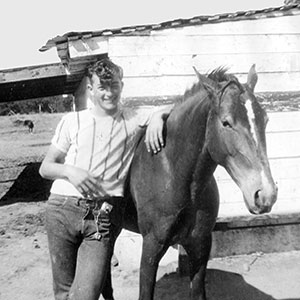 Black-and-white photo of a young man smiling with his arm around a horse, standing in front of a wooden building on a sunny day.