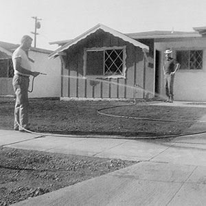 Two men stand in front of a mid-century house. One man waters the yard with a hose, while the other stands near the porch. The yard has bare dirt and there is a sidewalk in the foreground.