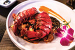 A plated dish featuring two lobster tails on a bed of vegetables, garnished with a purple orchid flower and a swipe of orange sauce on a white plate.