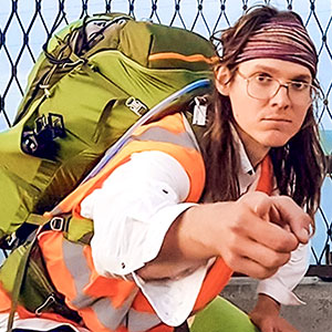 A person wearing glasses, a headband, and an orange safety vest crouches with a large green backpack, pointing towards the camera. A fence and blue sky are visible in the background.