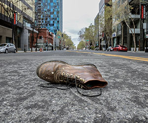A single worn brown boot lies abandoned in the middle of an empty city street, surrounded by tall buildings and trees on a cloudy day.