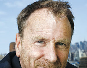 A middle-aged man with short brown hair and a trimmed beard smiles slightly while standing outdoors, with a city skyline blurred in the background under a blue sky.
