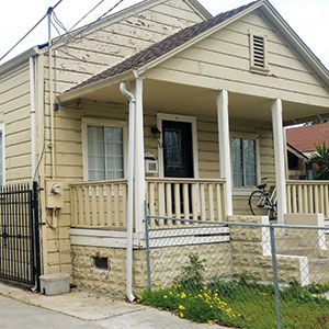 A small beige house with visible peeling paint, a covered front porch with white railings, a bicycle resting on the porch, and a metal fence bordering the yard with some yellow flowers near the walkway.