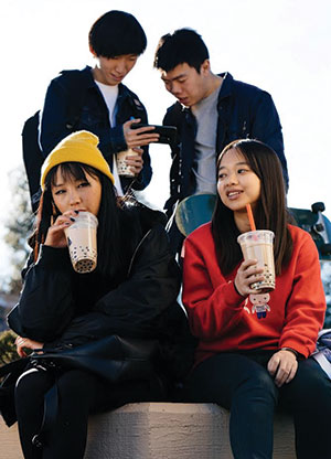 Two young women sit and drink bubble tea, one wearing a yellow beanie and the other a red sweater, while two young men stand behind them looking at a phone together.