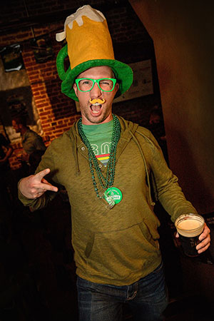 A man wearing a large beer mug hat, green glasses, and green beads smiles and makes a peace sign while holding a pint of dark beer. He is dressed festively, likely celebrating St. Patrick’s Day.