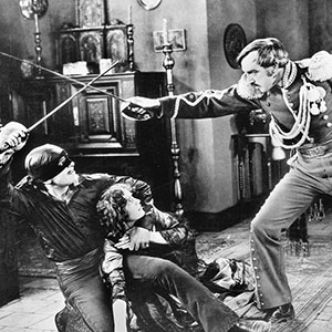 Three people are in a dramatic scene: a masked man with a sword duels a uniformed man, while a woman sits on the floor between them, appearing distressed, in an old black-and-white film setting.