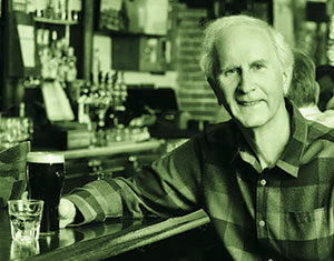 An older man in a plaid shirt sits at a bar, smiling at the camera, with a pint of dark beer and a glass of clear liquid in front of him. The background shows shelves and a person seated at the bar.