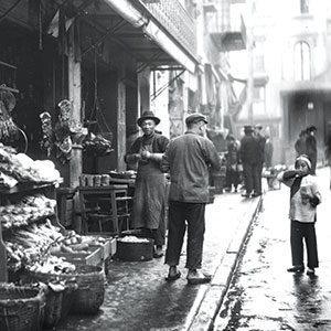 Black and white photo of a busy street market scene; vendors stand beside stalls with fruits and vegetables, while people walk and a child drinks from a cup in the foreground. Buildings line the narrow street.