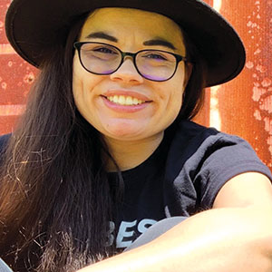 A person with long dark hair, wearing glasses, a black hat, and a black shirt, is smiling while sitting outdoors in front of a rust-colored corrugated metal wall.