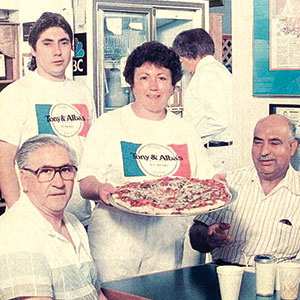 Four people are gathered in a pizzeria. A woman holds a pizza, standing next to a young man, while two older men sit at a table with drinks. All appear happy, and two wear white restaurant T-shirts. The scene is bright and casual.