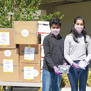 Two young people wearing masks and gloves stand beside stacked cardboard boxes labeled Face Shields outdoors, holding packaged face shields, with greenery and a building in the background.