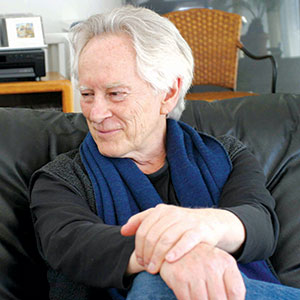 An older man with white hair sits on a black couch, smiling and looking to the side. He is wearing a dark shirt and a blue scarf, with his arms casually crossed over his knees.