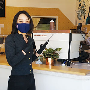 A woman wearing a dark mask and gloves stands behind a coffee counter, holding a metal wand near a white espresso machine. A small potted plant sits on the counter beside her.