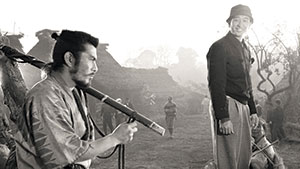 Two men stand in a rural village setting; one is sitting with a long stick over his shoulder, wearing traditional Japanese clothing, while the other stands smiling, wearing a hat and modern attire. Huts and trees are visible in the background.