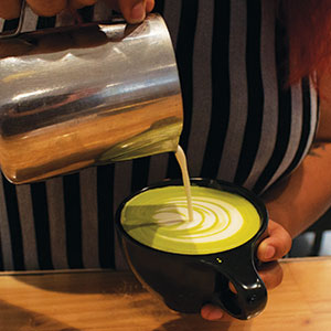A person pours steamed milk into a cup of green matcha latte, creating latte art. The person is wearing a black and white striped shirt.