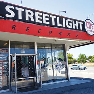 Streetlight Records storefront with large sign reading STREETLIGHT RECORDS CDS VIDEO. The entrance has metal bars, glass doors, and posters in the windows. A car and trees are visible on the street outside.