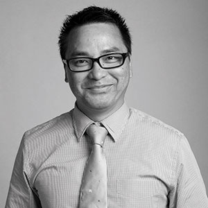 A man with short, dark hair and glasses smiles at the camera. He is wearing a light-colored, button-up shirt with a striped pattern and a light-colored tie. The background is plain and light.