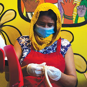 A woman wearing a yellow scarf, face mask, gloves, and red apron peels a banana in front of a colorful mural with hands and flowers on a yellow background.