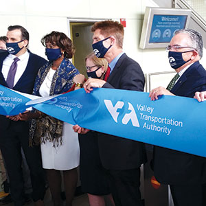 Six people wearing masks stand together holding and cutting a large blue ribbon that reads Valley Transportation Authority at an indoor ribbon-cutting ceremony.