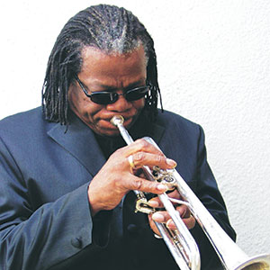 A man wearing sunglasses and a dark suit plays a trumpet. He has braided hair and is focused on the instrument, standing against a plain white background.