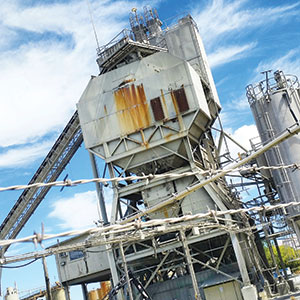 An industrial facility with large metal structures and conveyors, showing areas of rust and weathering, set against a bright blue sky with wispy clouds. Power lines run across the foreground.