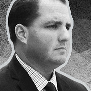 Black and white photo of a man in a suit and checkered shirt, looking serious and facing right, with a textured background.