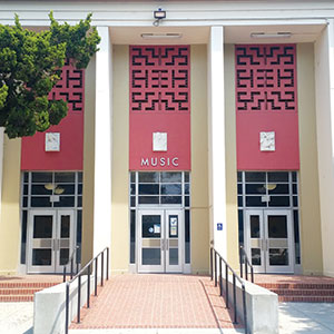 A building entrance with three large doors and “MUSIC” written above the center door. The facade has red decorative panels and a tree is partially visible on the left side. Steps and railings lead up to the entrance.