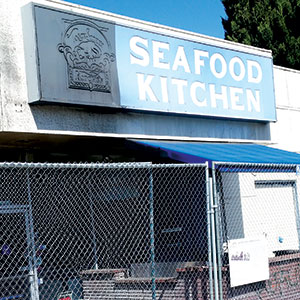 A sign reading Seafood Kitchen is mounted above a building entrance, which is partially obscured by a chain-link fence. The scene is outdoors and a blue awning is visible.