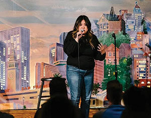 A woman stands on stage holding a microphone, performing stand-up comedy in front of an audience. The backdrop features colorful illustrations of city buildings.
