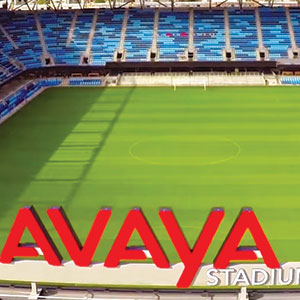 A soccer stadium with blue seats and a green field. Large red letters spelling AVAYA are prominently displayed in the foreground.