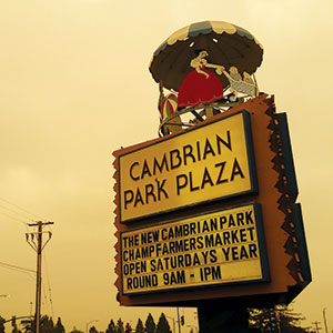 A vintage sign for Cambrian Park Plaza with a carousel horse and a couple on top. The sign advertises a farmers market open Saturdays year-round from 9 a.m. to 1 p.m. The sky is overcast.