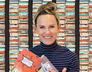A smiling woman in a striped turtleneck holds and points to two books in front of a colorful background made of stacked books.