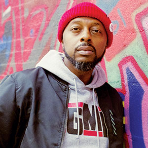 A man with a trimmed beard wears a red beanie, black jacket, and gray hoodie with lettering. He stands in front of a vibrant, colorful graffiti wall.