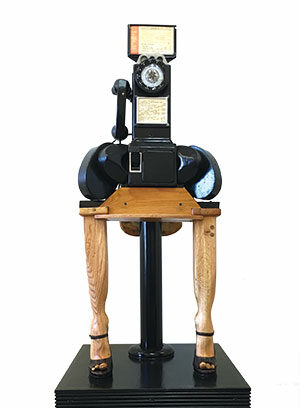 A sculpture resembling a vintage rotary telephone with handwritten notes, mounted on a wooden table with two legs shaped like animal legs, all standing on a black pedestal.