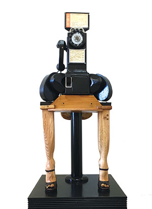 A sculpture resembling a vintage rotary telephone with handwritten notes, mounted on a wooden table with two legs shaped like animal legs, all standing on a black pedestal.