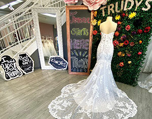 A white lace wedding gown with a long, intricate train is displayed on a mannequin in a bridal shop. Signs nearby read “I said Yes at Trudys.” Flowers and greenery decorate the background.