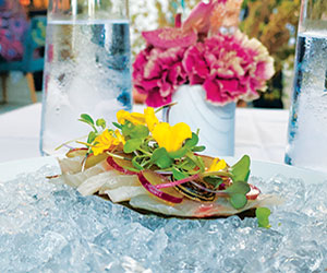 A plate of thinly sliced fish garnished with microgreens and yellow edible flowers is served on a bed of ice. Two tall glasses of water and a vase of pink flowers are in the background.
