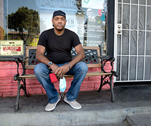 A man in a black t-shirt and blue jeans sits on a bench outside a building, holding a blue face mask. Signs and a metal security door are visible behind him.