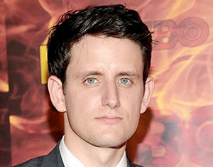 A man with short dark hair wearing a gray tuxedo and black bow tie stands in front of a backdrop with a fiery orange and yellow pattern and the HBO logo.