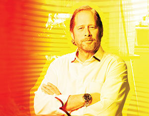 A man with light hair and a beard stands with arms crossed, wearing a white shirt and wristwatch. The image has a warm, yellow and red overlay with abstract lines in the background.