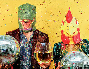 Two people wearing dinosaur and chicken masks hold disco balls and a large wine glass, standing in front of a yellow background with colorful confetti falling around them.
