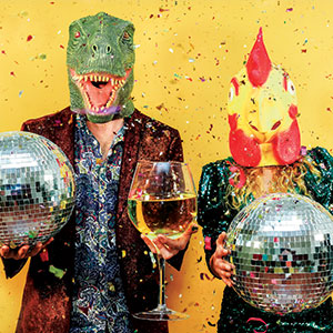 Two people wearing dinosaur and chicken masks hold disco balls and a large wine glass, standing in front of a yellow background with colorful confetti falling around them.