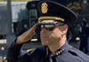 A police officer in uniform and sunglasses salutes, with other officers and a police vehicle visible in the background.