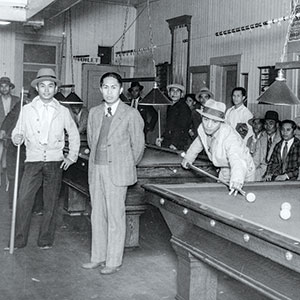 Black and white photo of several men in hats and suits gathered around pool tables in a billiards hall, with some watching and one man preparing to take a shot.