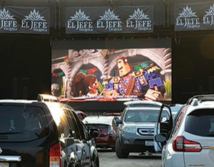 Cars parked at a drive-in theater watch an animated movie on a large outdoor screen, with sunlight reflecting off the vehicles and El Jefe Tequila banners above the screen.